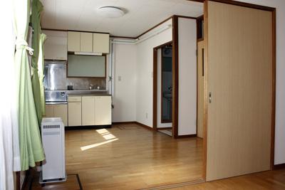 正面にキッチン、床は茶色いフローリンクで白い壁紙、左手前にストーブが設置されている室内写真