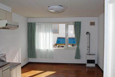 正面に窓があり、茶色のフローリングの床で白い壁紙、左手にキッチンがある室内写真