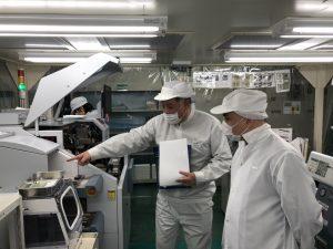 株式会社京都セミコンダクター上砂川事業所：帽子とマスク、白い作業着で機械が並ぶ中、説明を受ける男性の写真